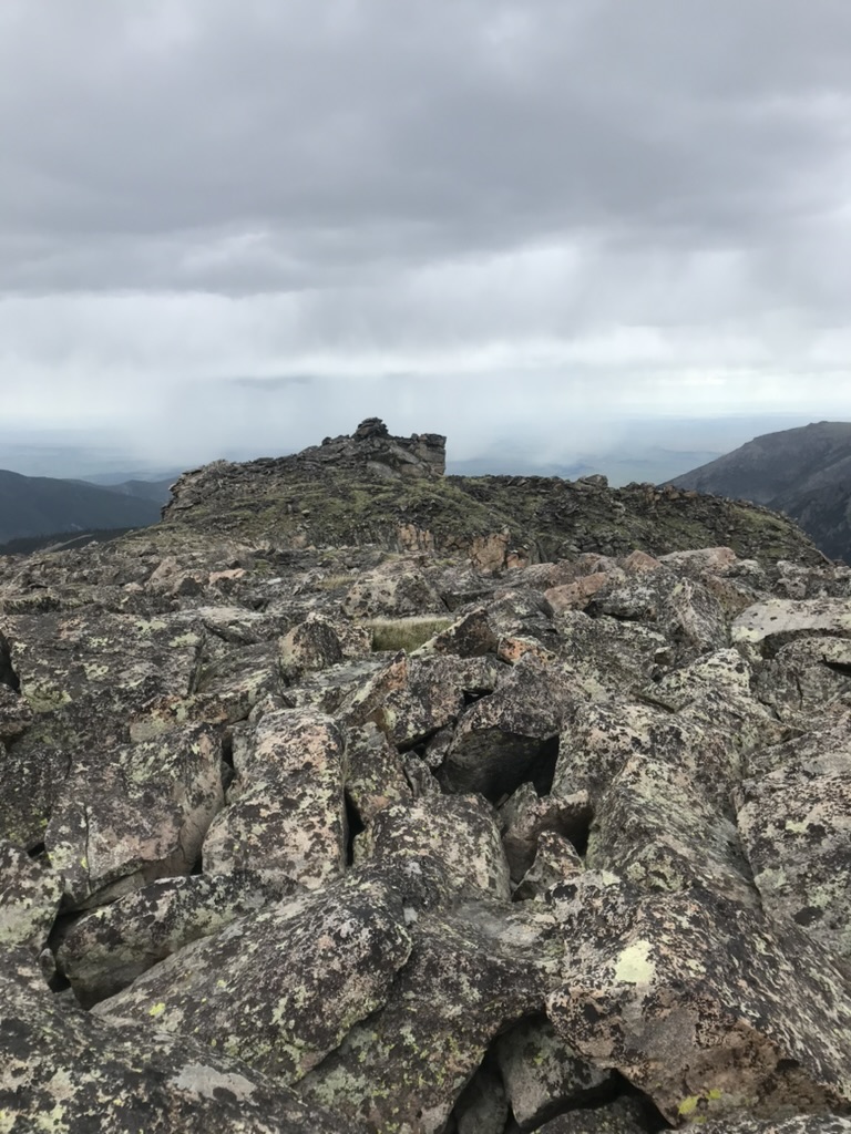 A lichen-covered rocky summit under a gray sky, with distant ranges fading into weather.